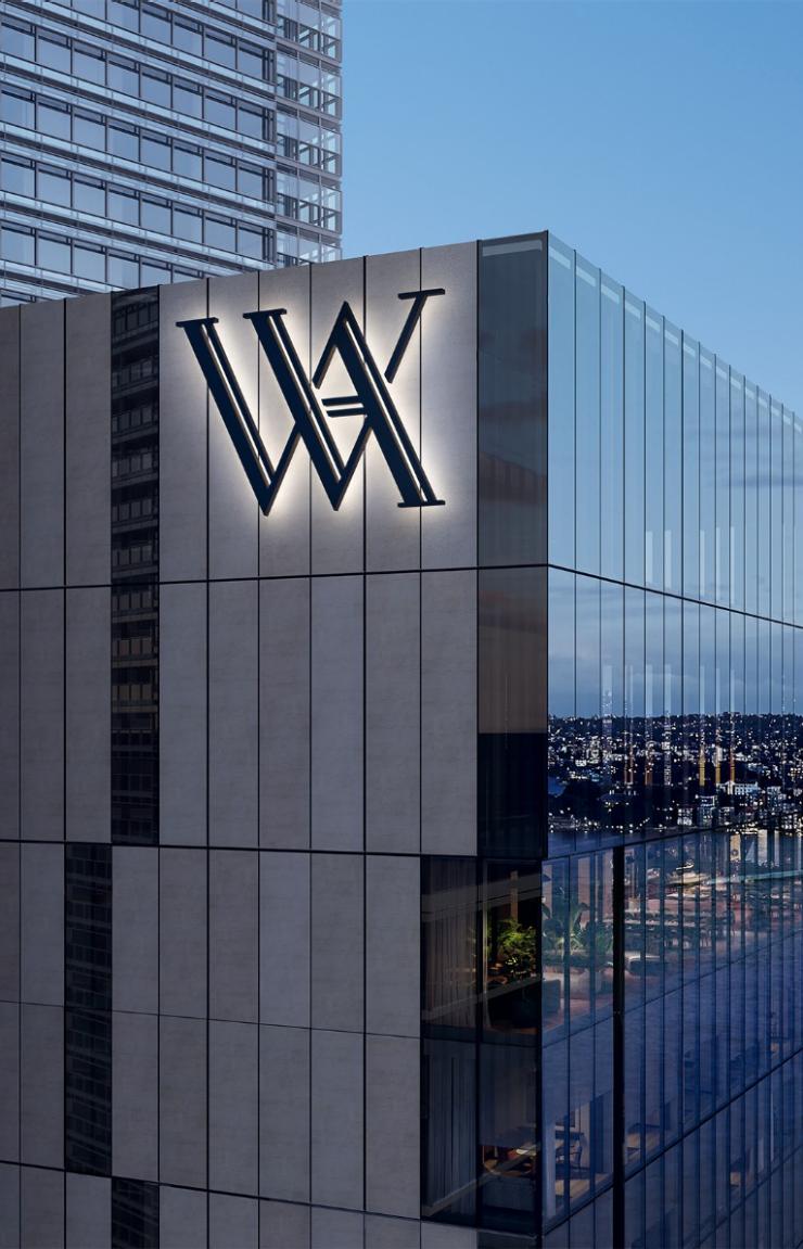 A sleek skyscraper bearing an illuminated WA emblem looks out over a glowing harbour, bridge, and Opera House at dusk in Sydney, New South Wales. © Hilton