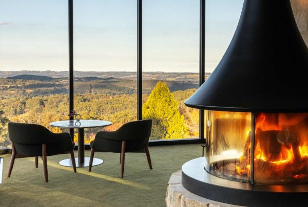 A crackling stone-encased circular fireplace warms a lounge with floor-to-ceiling windows revealing sweeping views of the Adelaide Hills, Sequoia Lodge, Adelaide, South Australia. © Sequoia Lounge