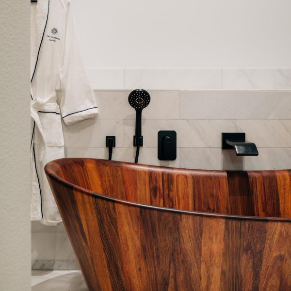 A luxurious timber bathtub stands beside matte black fixtures and a plush white bathrobe hanging against marble wall tiles, The Tasman, Hobart, Tasmania. © The Tasman