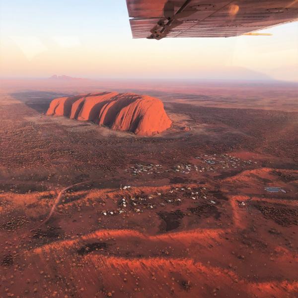 An aerial view framed by an airplane wing shows Uluru bathed in warm sunlight above the red desert landscape, Uluru-Kata Tjuta National Park, Northern Territory. © Tourism Australia