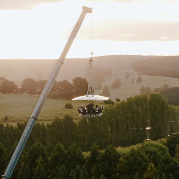 A crane suspends a platform of diners high above rolling green hills at sunset during a Dinner in the Sky experience. © Dinner in the Sky