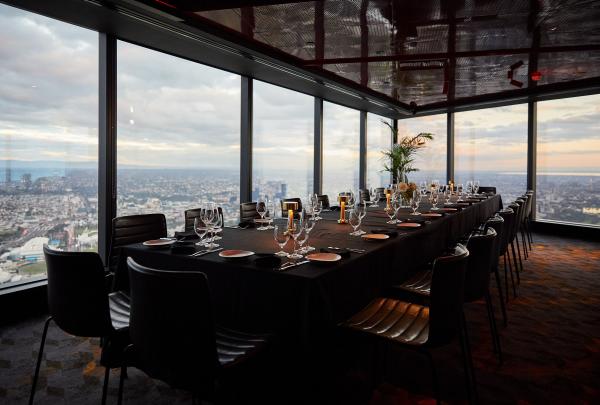 A long dining table set with glassware inside a high-rise room with floor-to-ceiling windows offering expansive city views, Eureka Tower, Melbourne, Victoria. © Tourism Australia