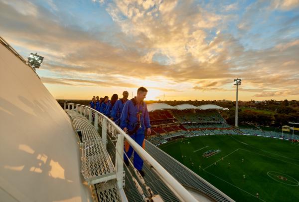 Guests in safety harnesses walk along a high roof platform overlooking a stadium field at sunset, RoofClimb, Adelaide Oval, Adelaide, South Australia. © Adelaide Oval