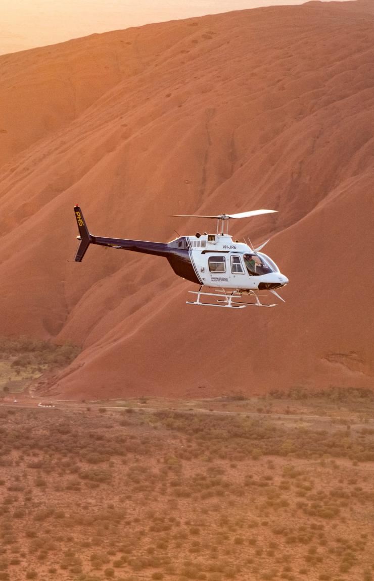A helicopter flies past the glowing red sandstone rock face of Uluru in the early morning light, Uluru-Kata Tjuta National Park, Northern Territory. © Tourism NT