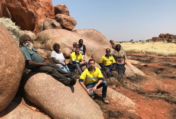 Mark Savich and Tom Lyons sit alongside Traditional Owners from the Kiwirrkurra community on large red rock boulders in an arid outback landscape in Western Australia. © Agrimin
