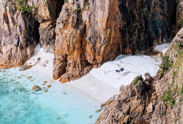 A helicopter rests on a secluded white sand beach nestled between steep rocky cliffs and turquoise ocean waters, Hayman Islands, Queensland. © Tourism Australia