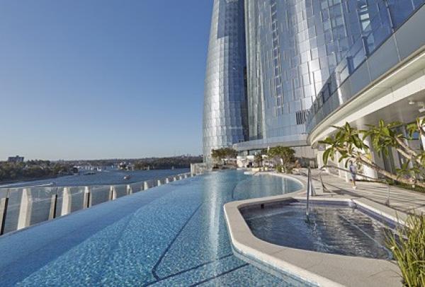 An infinity pool and spa tub stretch along an outdoor deck beneath the soaring Crown Sydney skyscraper, overlooking Sydney Harbour, New South Wales. © Crown Sydney