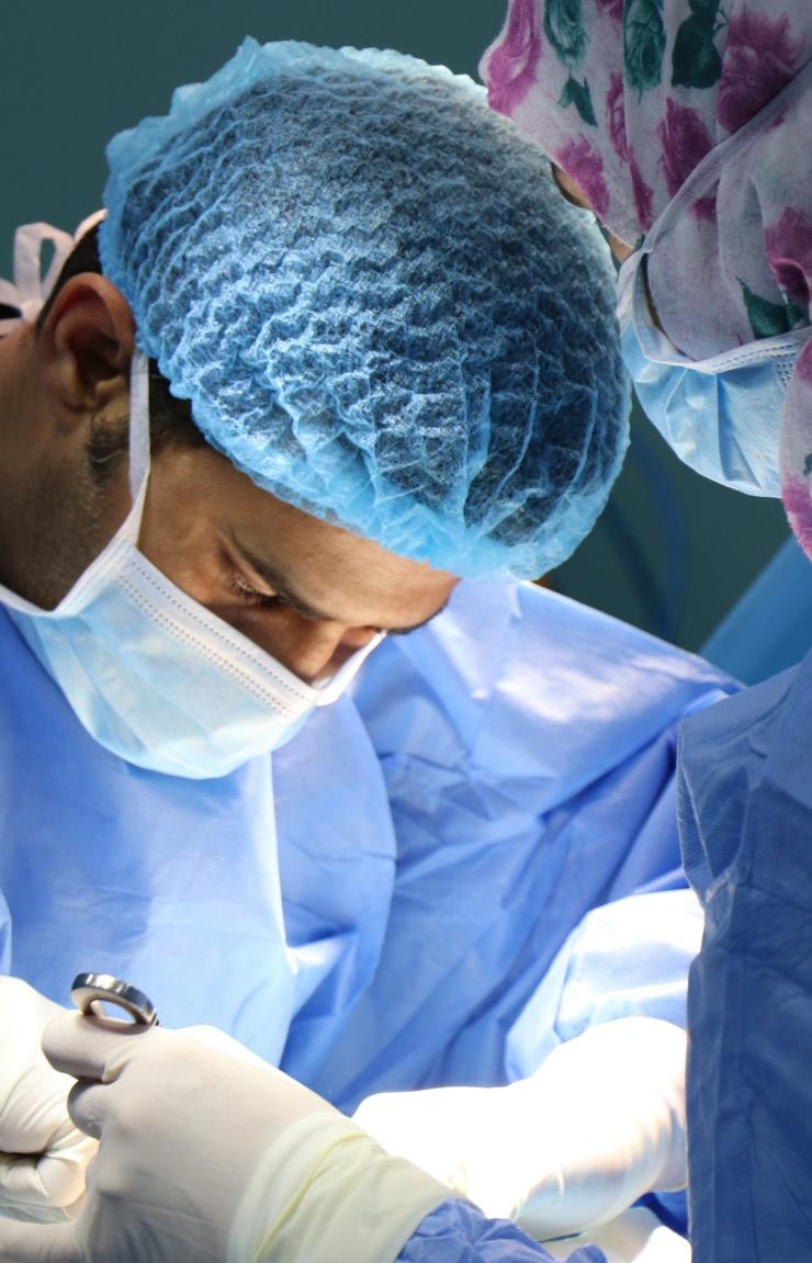 Dr Geoff Rogers wears blue scrubs and a surgical mask while performing a precise medical procedure. He focuses intently on his hands as he uses specialised tools, assisted by a colleague in a floral head cover. © Jafar Ahmed