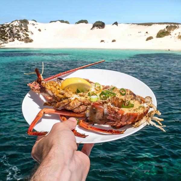 A hand holding a plate with a freshly cooked rock lobster dish above clear turquoise water during a Rottnest Cruises tour on Rottnest Island, Western Australia. © Rottnest Cruises