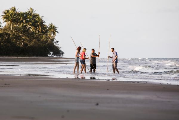 A group stands in shallow ocean water at the shore holding wooden spears, learning traditional spearfishing techniques on a Walkabout Cultural Adventures tour in Tropical North Queensland. © Tourism Australia