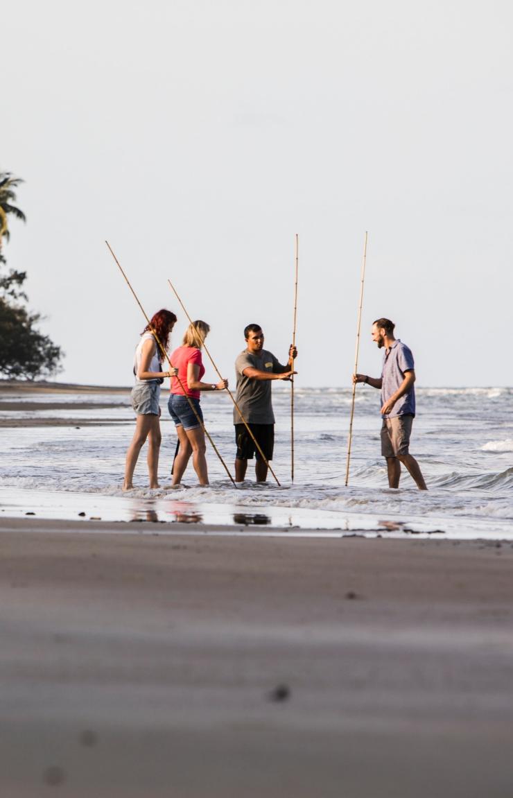 A group stands in shallow ocean water at the shore holding wooden spears, learning traditional spearfishing techniques on a Walkabout Cultural Adventures tour in Tropical North Queensland. © Tourism Australia