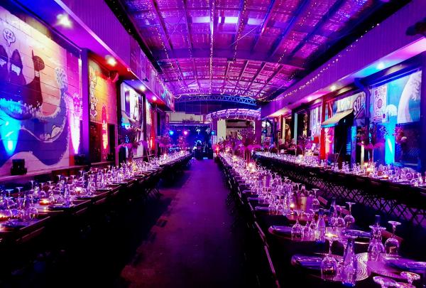 Long banquet tables set with glassware stretch down a colourful lane illuminated by vibrant purple overhead lighting at Miami Marketta, Gold Coast, Queensland. © Tourism Australia