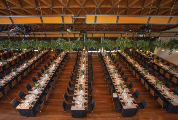 Long, parallel dining tables adorned with hanging greenery fill a spacious wooden hall at National Wine Centre of Australia, Adelaide, South Australia. © Tourism Australia