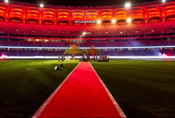 A red carpet leading across the green grass toward an exclusive Optus Stadium Dinner on the Ground event under bright stadium lights at night in Perth, Western Australia. © Tourism Australia