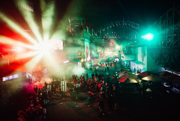 Vibrant crowds gather under colourful laser light displays at Luna Park during a night event, with the Sydney Harbour Bridge looming in the background in Sydney, New South Wales. © Luna Park