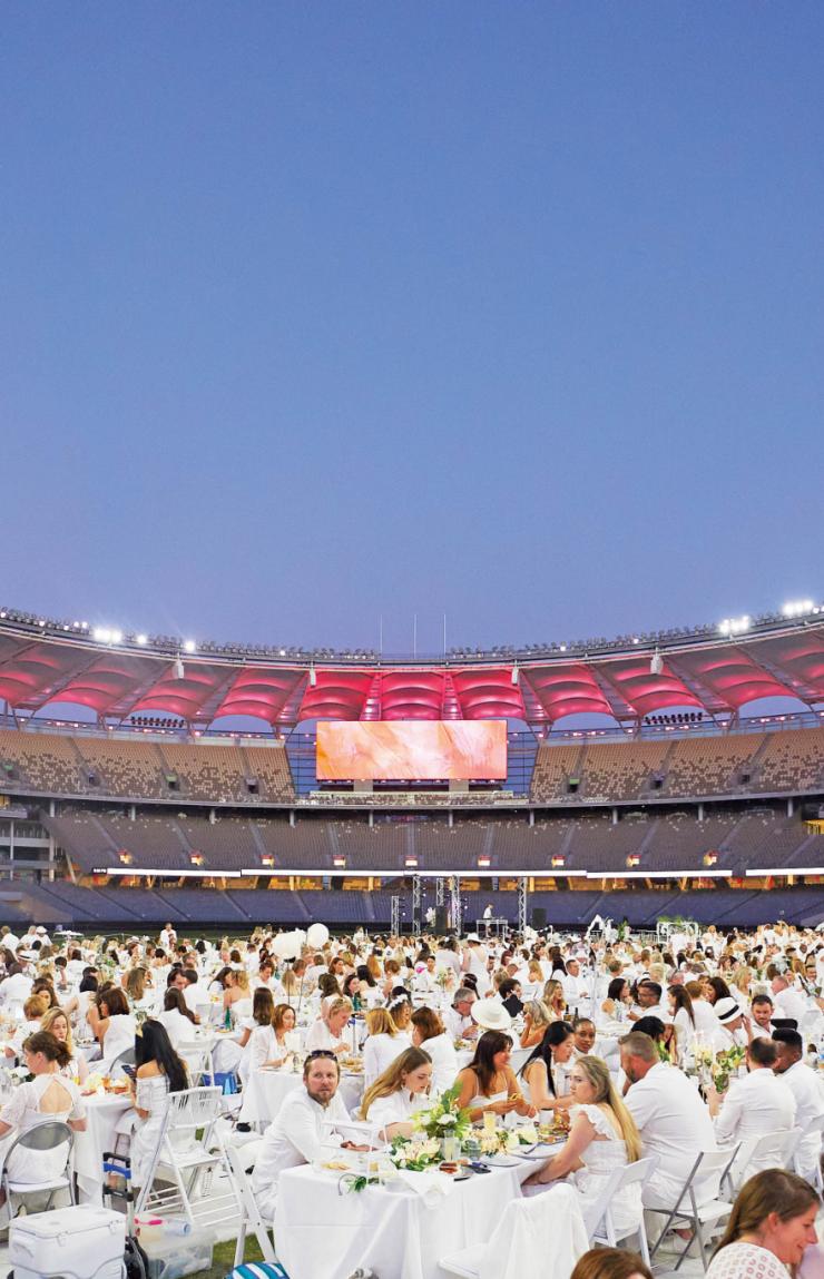Guests dressed in all white attend an outdoor dining event on the illuminated field inside Optus Stadium at dusk in Perth, Western Australia. © Tourism Australia