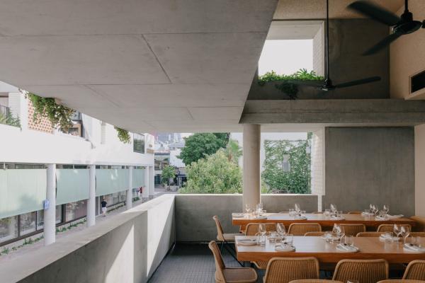 An open-air private dining room features wooden tables and woven chairs along a sleek concrete balcony looking onto street greenery, sAme sAme, The Calile Hotel, Brisbane, Queensland.© The Calile Hotel