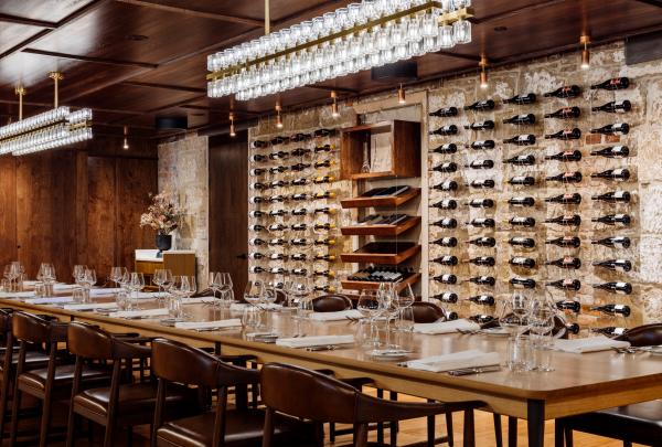 A long timber table set with wine glasses sits beneath modern linear lights against a exposed sandstone wall filled with wine racks, The Printing Room, Peppina, Hobart, Tasmania. © Peppina
