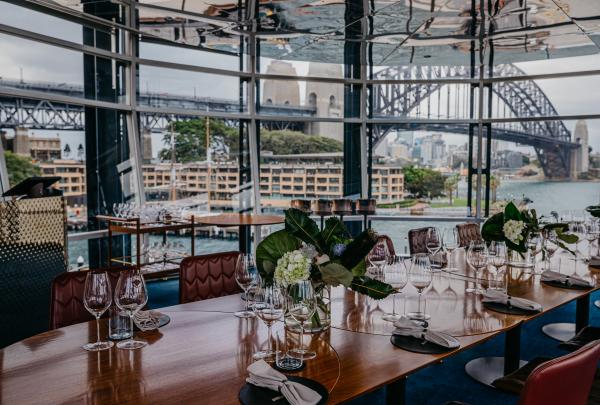 An elegantly set long dining table with wine glasses offers sweeping views of the Sydney Harbour Bridge through floor-to-ceiling windows, Upper Tower, Quay, Sydney, New South Wales. © Quay