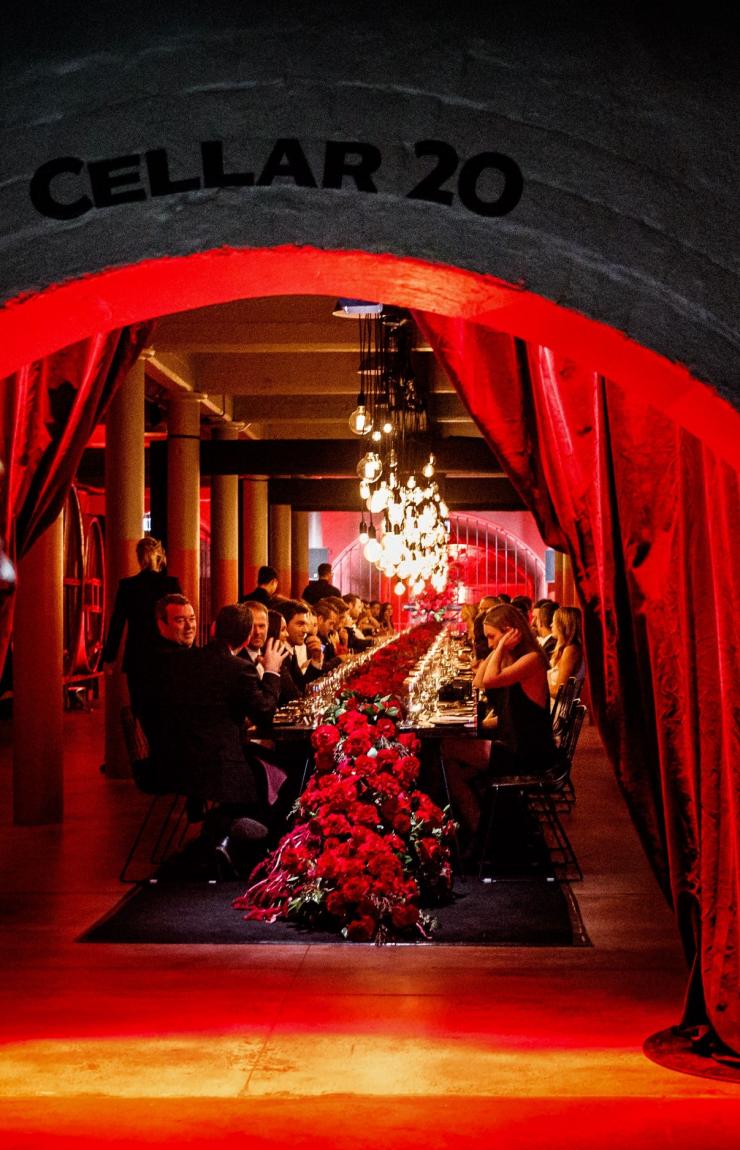 Guests dine in a dramatic, red-lit underground wine cellar decorated with vibrant floral displays, Penfolds Dining Room, Barossa, South Australia. © Tourism Australia