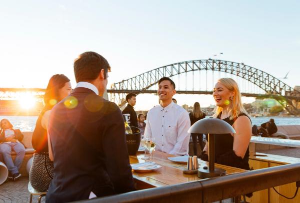 Friends socialise with drinks at an outdoor harbourfront table at Opera Bar as sunset casts a warm golden glow over the Sydney Harbour Bridge in Sydney, New South Wales. © Destination NSW