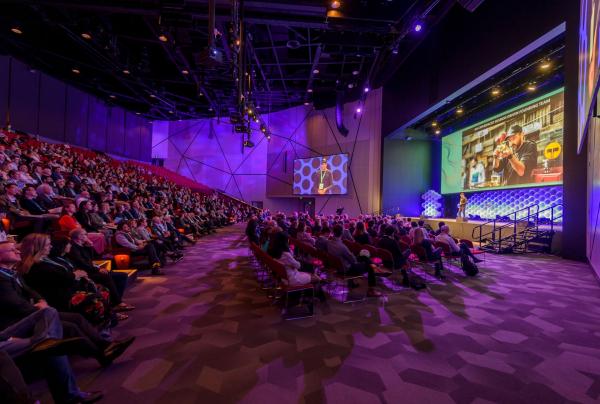 An audience fills a modern, purple-lit auditorium while watching a speaker present on stage near large screen displays at the Ozwater'21 Conference in Brisbane, Queensland. © Simon Casson
