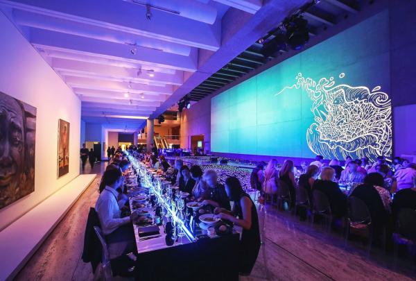 Guests enjoy a formal dinner at long banquet tables in a gallery hall illuminated by blue lighting and vibrant projected wall projections at the Dreamtime 2017 event in Brisbane, Queensland. © Remco Jansen