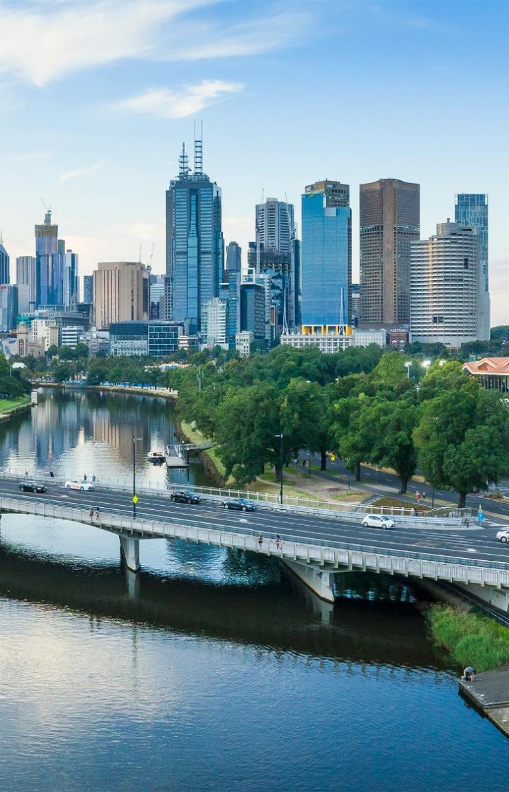 An aerial view shows the tranquil Yarra River flowing beneath a bridge towards the towering city skyline and sports precinct of Melbourne, Victoria. © Visit Victoria