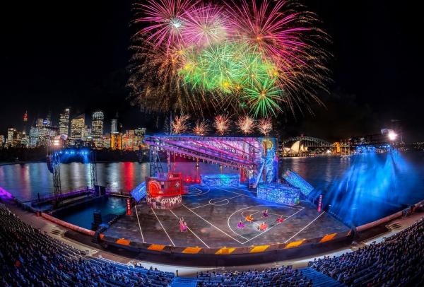 Fireworks burst over a vibrant floating outdoor stage set for Handa Opera on Sydney Harbour, with the illuminated city skyline and Opera House in the background, Sydney, New South Wales. © Opera Australia