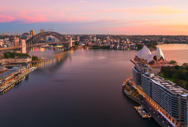 An aerial sunset view over Sydney Harbour and Circular Quay, showing the Sydney Opera House and Sydney Harbour Bridge bathed in pink sky, Sydney, New South Wales. © Destination NSW