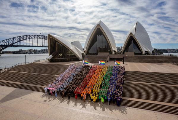 A large crowd dressed in pride colours forms a human progress flag on the steps of the Sydney Opera House for WorldPride in Sydney, New South Wales. © Daniel Boud