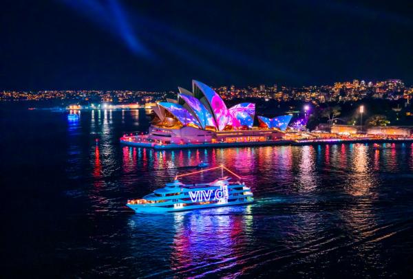 The Sydney Opera House glows with colourful light projections as a illuminated boat cruises across the dark harbor during Vivid Sydney in Sydney, New South Wales.© Destination NSW