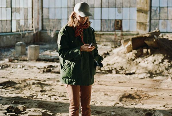 Wearing a cap and green jacket, a woman checks her phone inside a sunlit, ruined warehouse filled with rubble, captured on set for a music video with Aurora. © Kristof Brandl