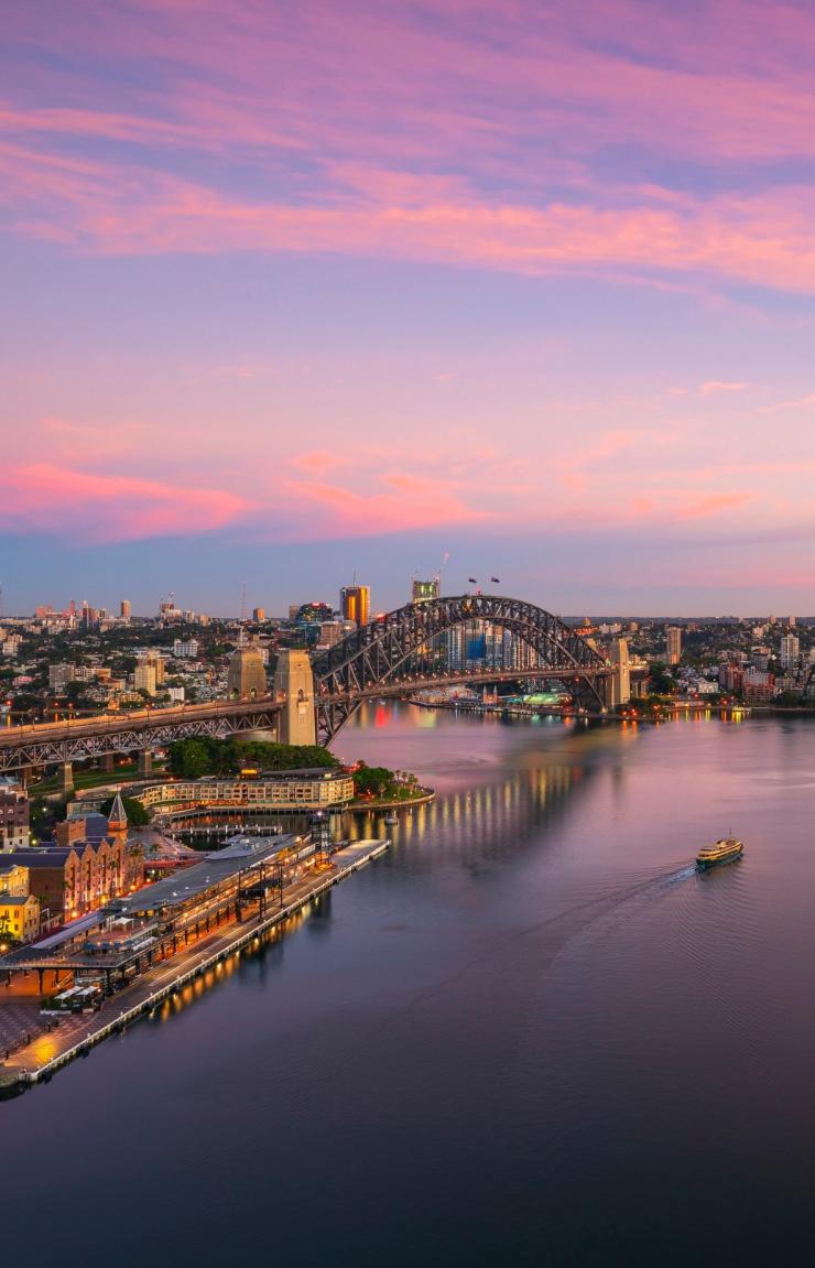 An aerial view of the Sydney Harbour Bridge and Sydney Opera House set against vibrant pink sunset skies reflecting over Sydney Harbour, Sydney, New South Wales. © Destination NSW