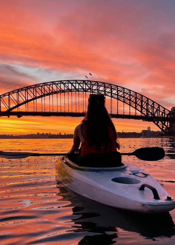 A kayaker paddles across calm waters toward the silhouetted Sydney Harbour Bridge beneath a fiery orange sunrise sky, Sydney Harbour, New South Wales. © Sydney by Kayak