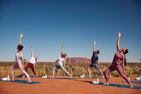 Visitors stretch in outdoor yoga poses on mats set against a vibrant red desert landscape with the iconic Uluru in the distance, Uluru-Kata Tjuta National Park, Northern Territory. © NT Business Events