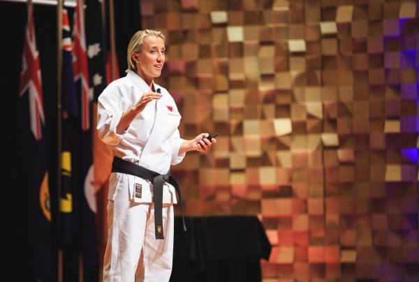 Speaker, Nadine Champion, wears a white martial arts uniform and black belt, gesturing on stage beside Australia flags. © Saxton Speakers