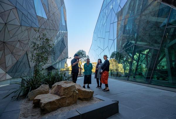Visitors gather outdoors between striking modern geometric glass structures to hear a First Nations guide speak during an experience with Koorie Heritage Trust, Melbourne, Victoria. © Tourism Australia