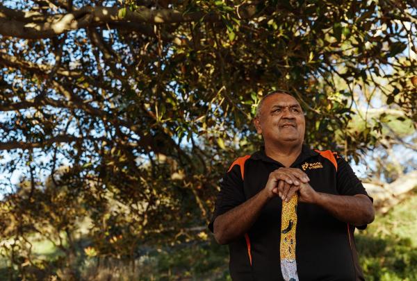 First Nations guide holding a painted clapstick rests under dappled sunlight beneath leafy branches during a Wuldi Cultural Tour, hosted at Gemtree Wines, South Australia. © SATC