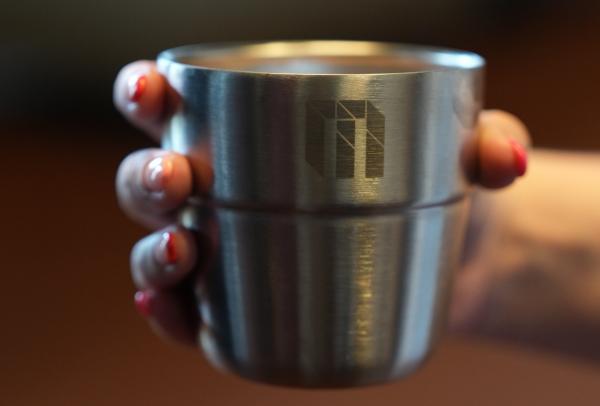 A hand with painted fingernails holds a stainless steel cup emblazoned with a modern logo at the Melbourne Convention and Exhibition Centre, Melbourne, Victoria. © Mat Vendeville