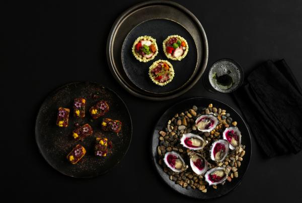 An overhead view of gourmet canapés, tartlets, and fresh oysters neatly arranged on dark plates against a black surface, catering created by ICC Sydney, Sydney, New South Wales. © ICC Sydney