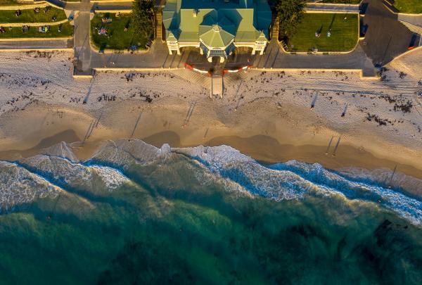 An aerial view of gentle surf breaking onto golden sand alongside the iconic Indiana Teahouse and green terraced lawns at Cottesloe Beach, Perth, Western Australia. © Tourism Australia