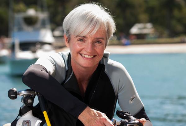 Professor Emma Johnston smiles while wearing a wetsuit and holding scuba diving gear on a sunny wharf beside clear blue water at Sydney Harbour, Sydney, New South Wales. © UNSW Sydney