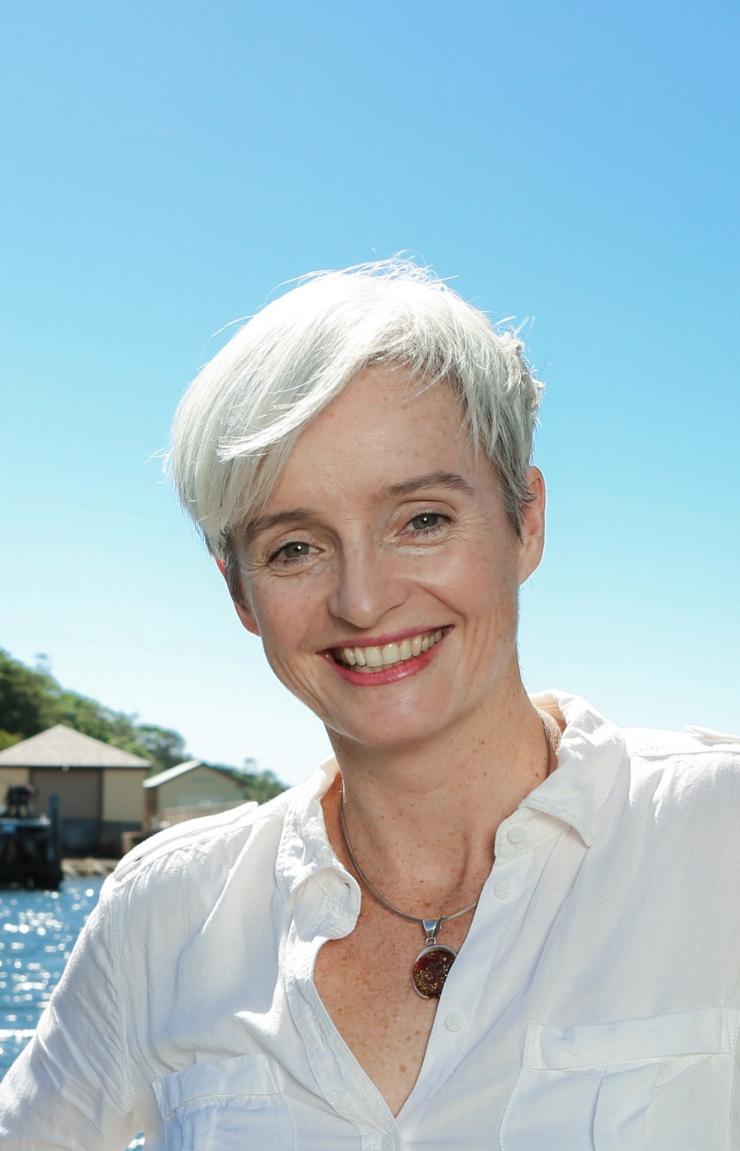 Professor Emma Johnston smiles outdoors against a sparkling blue waterway and lush coastal hillside. © UNSW Sydney