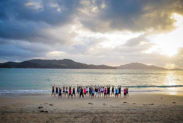 A group stretches with arms raised during outdoor yoga on the beach shore of Hamilton Island in the Whitsundays, Queensland, under a dramatic sky at sunrise with ONeill Photographics capturing it. © ONeill Photographics