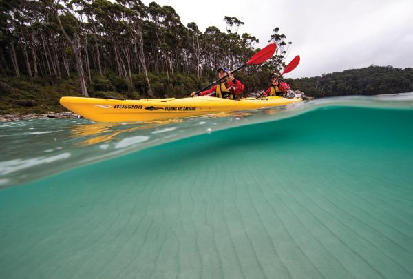 Two paddlers glide across crystal-clear turquoise waters in a bright yellow tandem kayak with Roaring 40s Kayaking, framed by a forested Tasmanian shoreline along the glass-like water surface. © Roaring 40s Kayaking