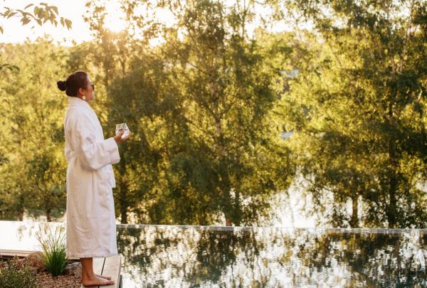 A woman in a white bathrobe holds a drink while relaxing poolside amidst lush green trees illuminated by afternoon light at Lake House Daylesford, Victoria. © Lake House Daylesford