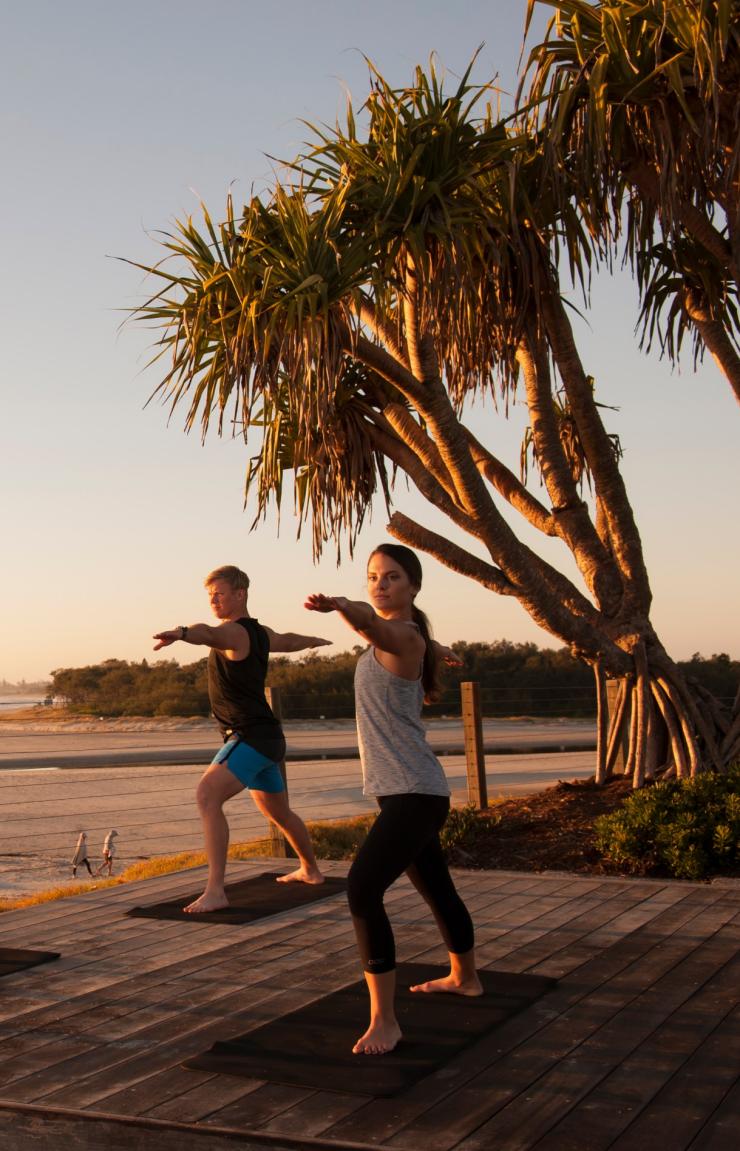 Three people practice Sunrise Yoga on a wooden deck by the beach, holding warrior poses under glowing morning light at Elements of Byron Bay, Byron Bay, NSW. © Destination NSW