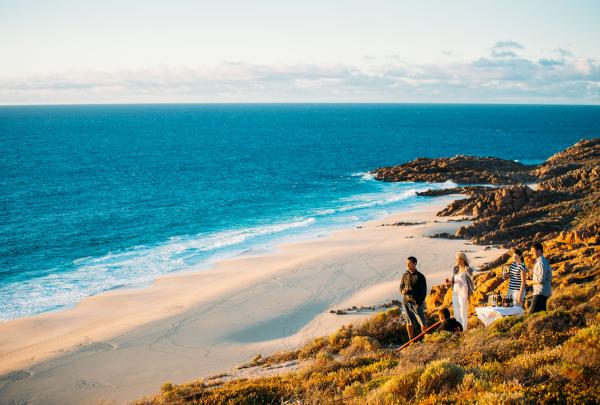 A small group enjoys wine and drinks atop coastal cliffs overlooking a pristine sandy beach and turquoise ocean during golden hour, Margaret River region, Western Australia. © Mark Boskell