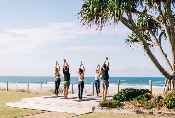 Guests practice yoga on an outdoor wooden platform overlooking a sunny beach under pandanus palm trees, Elements of Byron Bay, Byron Bay, New South Wales. © Paul Bramford